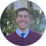 White male with dark brown hair has a big smile while standing outside of a building wearing a maroon sweater and blue button down with a dark blue tie.
