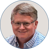 The speaker has light brown hair and is smiling while wearing a checkered blue button down shirt and black-rimmed glasses.