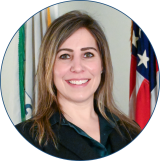 Erin has long brown and blond hair and is smiling while wearing a black blazer and standing in front of the US flag and the EPA flag.