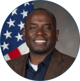 Profile picture of Harper wearing a brown blazer and dark blue button down while standing in front of the American flag.